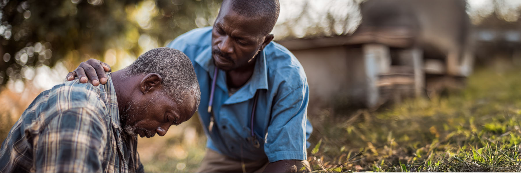 A compassionate outdoor scene showing one man comforting another. The man in a blue shirt, possibly a healthcare worker or minister, gently places his hand on the shoulder of a distressed man who is seated and bowing his head. The background is softly blurred with hints of nature and a rural setting, conveying empathy, support, and human connection.