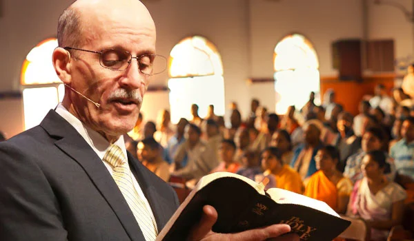 Pastor Doug wearing a headset microphone reads from the Holy Bible to a large, attentive congregation seated in a well-lit church, suggesting a scene of preaching or teaching during a religious service.