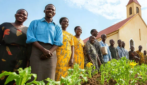 A group of smiling people standing together in front of a church building, behind a lush garden with green plants and yellow flowers, symbolizing community, joy, and agricultural growth.