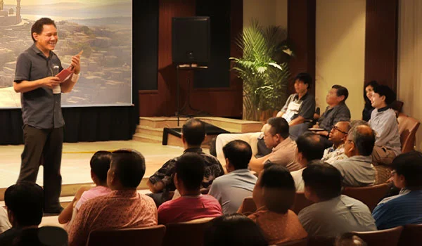 تظهر الصورة متحدثًا يخاطب جمهورًا في ما يبدو أنه ندوة أو جلسة تعليمية. الأجواء مريحة وجذابة - الناس جالسون بانتباه بينما المتحدث، ممسكاً بملاحظات أو كتاب، مبتسماً ويبدو أنه في منتصف عرض أو محاضرة. يتم إعداد شاشة عرض ونظام مكبر صوت، ويوحي الإعداد بمكان داخلي مثل قاعة مؤتمرات أو قاعة صغيرة.