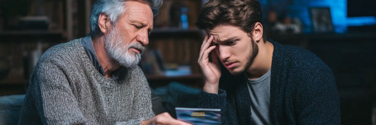 An older man and a younger man sitting closely together indoors, both looking concerned as they examine a document. The older man has gray hair and a beard, wearing a gray sweater, while the younger man looks distressed, resting his hand on his forehead and wearing a dark cardigan.