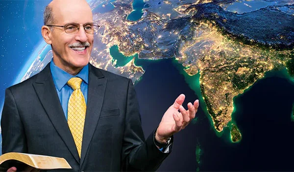 Pastor Doug holding an open Bible, gesturing as if speaking, with a satellite image of South Asia illuminated at night in the background, symbolizing global outreach or missionary work.