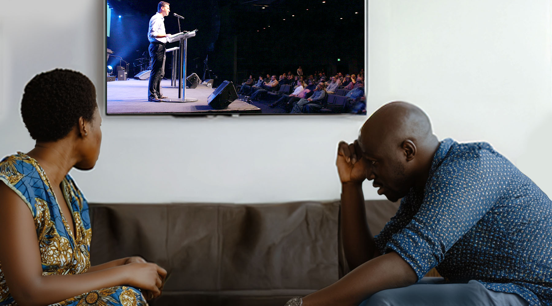 A couple sits on a couch in a living room watching a TV mounted on the wall. On the screen, a man stands at a podium speaking to a seated audience in a large auditorium. In the foreground, the woman looks toward her partner, while the man leans forward with his hand to his forehead, appearing worried or deep in thought.