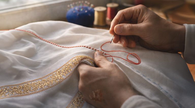 Close-up of a person’s hands carefully sewing red thread into white fabric trimmed with ornate gold embroidery, illustrating the themes “Clothed by Christ” and “Dressing for success.” A needle pulls bright red thread through the cloth along a neat seam with visible blue guide stitches, while spools of thread and a blue pin cushion rest softly blurred in the background near a window, highlighting detailed craftsmanship and intentional design.