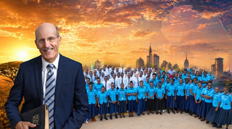 A smiling older man in a suit and striped tie holds a Bible in the foreground, standing against a vivid orange sunset sky. Behind him, a large group of students and adults in coordinated blue uniforms pose together outdoors, with a city skyline and trees visible in the background.