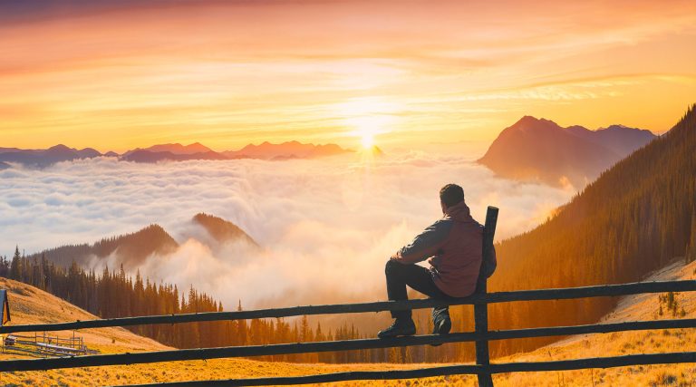 A man sits on a wooden fence overlooking a mountain landscape at sunrise, with golden light illuminating rolling hills, pine forests, and a sea of clouds below, while a small cabin rests on a grassy slope nearby.