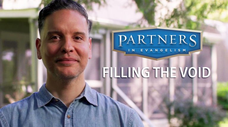 A man in a light blue button‑up shirt stands outdoors in front of a modern building with large windows and surrounding greenery. To his right is a blue and gold circular logo that reads “Partners in Evangelism” with the tagline “Filling the Void.”